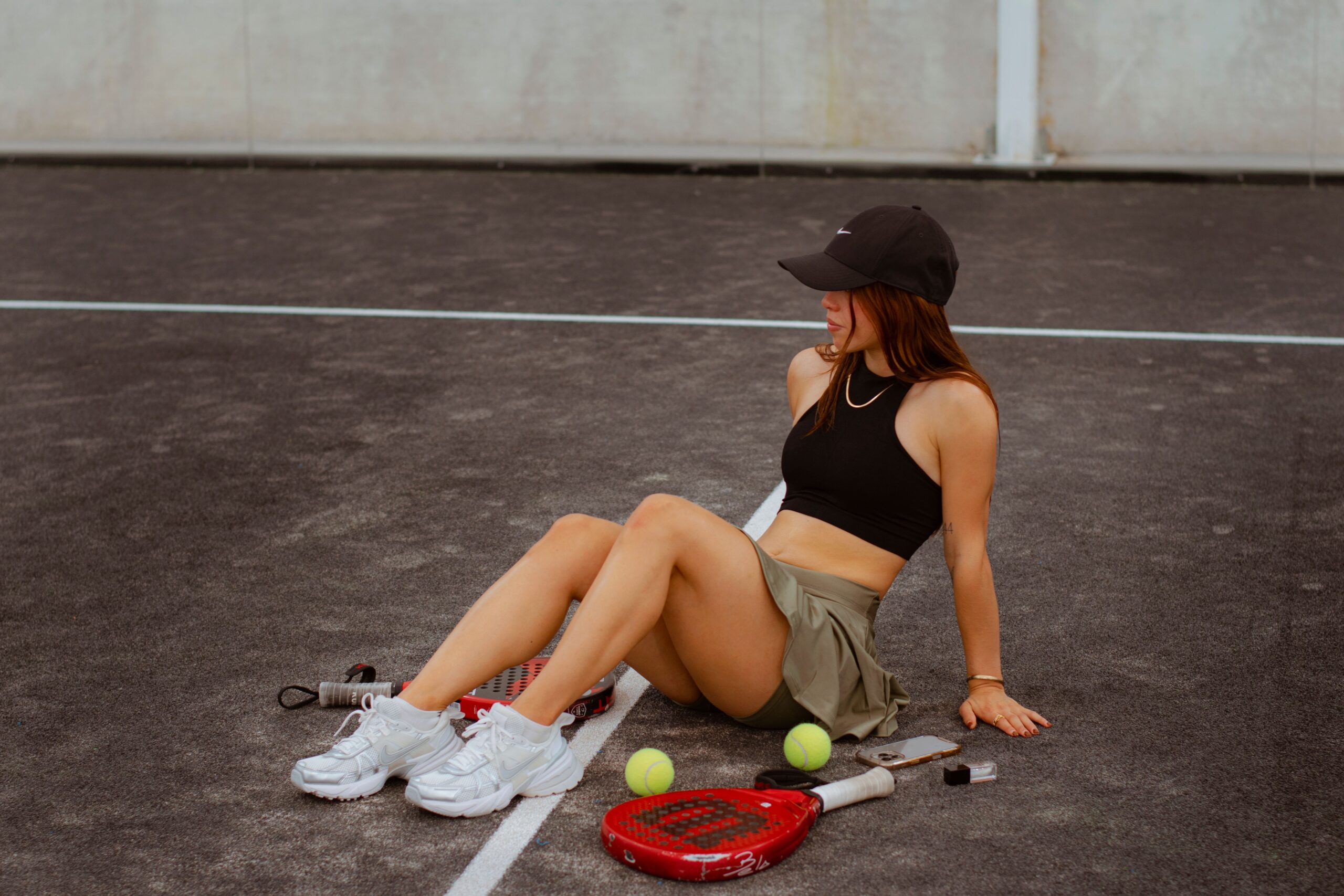 Woman relaxing on an outdoor tennis court wearing a black sleeveless crop top and khaki tennis skirt with tennis rackets and balls beside her.